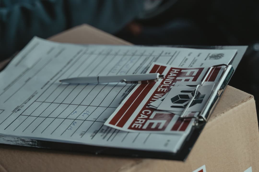 A shipping manifest and handling labels clipped to a cardboard box, illustrating the HS code vs HTS code vs Schedule B paperwork trail