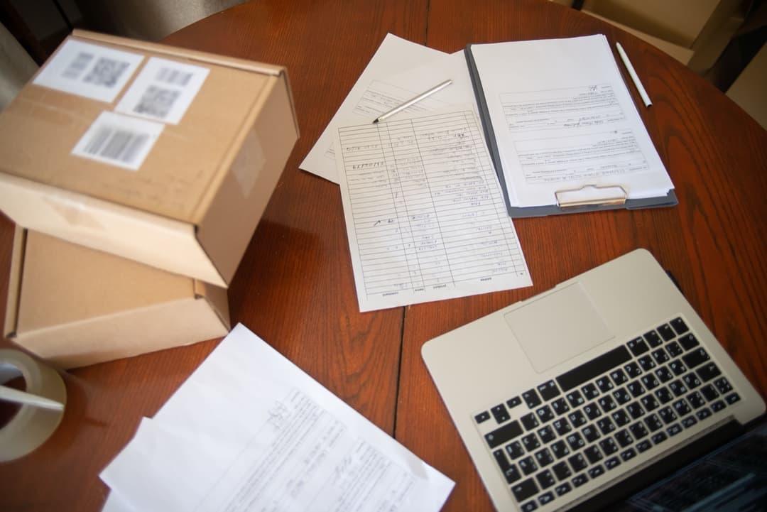 Invoice paperwork and shipping cartons on a desk, representing a landed cost calculation for an import shipment